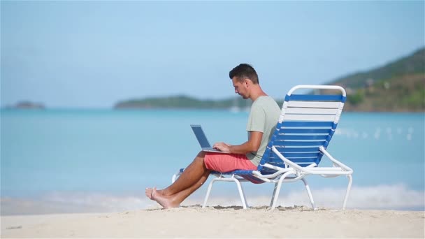 Young man with laptop on tropical caribbean beach. Man sitting on the sunbed with computer and working on the beach — Stock Video