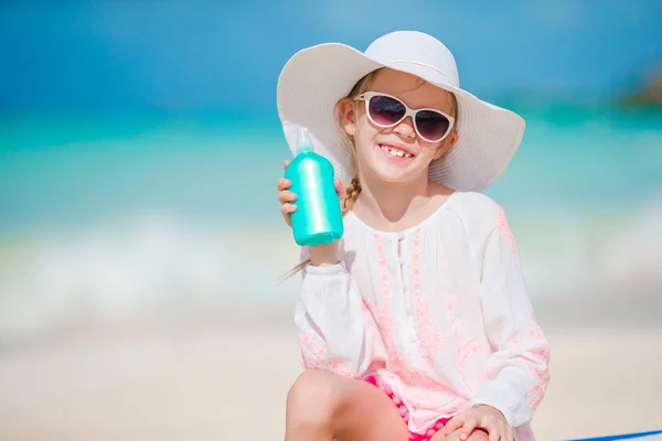 Little girl in hat with bottle of sun cream sitting at sunbed on tropical beach — Stock Photo, Image