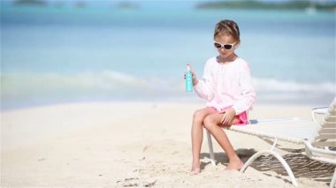 Tropikal plajda oturan güneş kremi şişeli küçük kız.
