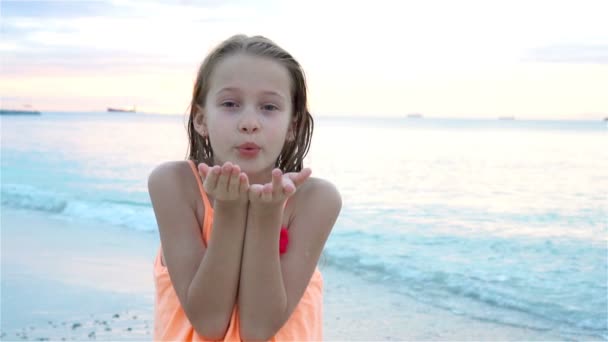 Roztomilá holčička na pláži se při západu slunce dobře baví. Šťastné dítě při pohledu na kameru a líbání pozadí krásné nebe a moře. SLOW MOTION — Stock video