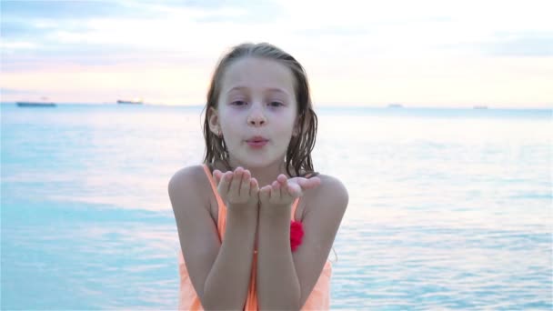 Schattig klein meisje op het strand heeft veel plezier bij zonsondergang. Gelukkig kind kijkend naar camera en kussende achtergrond prachtige lucht en zee. SLOW MOTION — Stockvideo