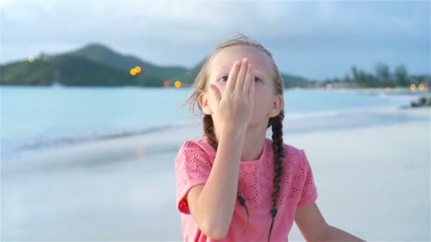 Liebenswertes kleines Mädchen am Strand, das bei Sonnenuntergang viel Spaß hat. Glückliches Kind schaut in die Kamera und küsst Hintergrund schönen Himmel und Meer. Langsame Motorisierung — Stockvideo