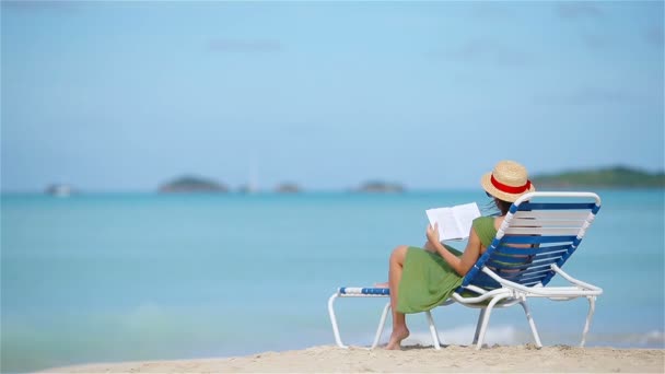Молодая женщина читает книгу о шезлонге на тропическом белом пляже — стоковое видео