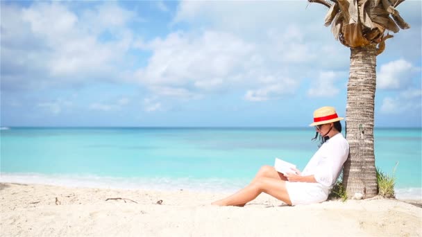 Mujer joven leyendo libro bajo la palmera en la playa — Vídeo de stock