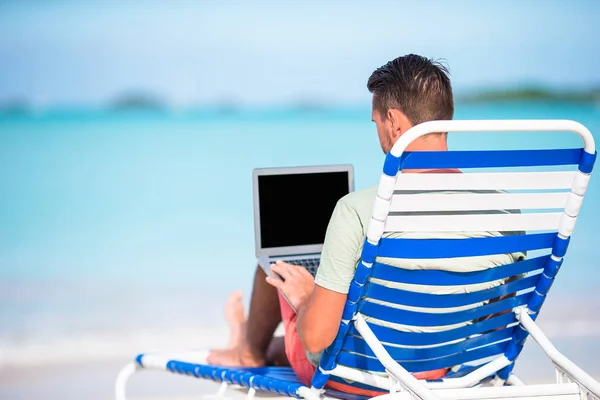 Young man with laptop on tropical caribbean beach. Man sitting on the sunbed with computer and working on the beach — Stock Photo, Image