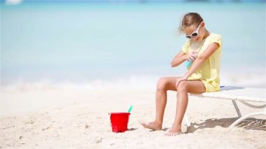 Tropik sahilde oturup güneş kremi şişe şapkalı küçük kız