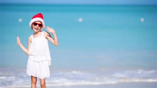 Kleines entzückendes Mädchen mit Weihnachtsmütze am weißen Strand während der Weihnachtsferien. glücklich lächelndes Kind blickt in die Kamera — Stockvideo