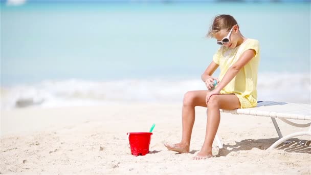 Liten flicka i hatt med flaska solkräm som sitter på tropical beach — Stockvideo