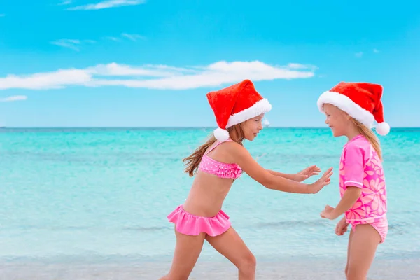 Las niñas adorables en los sombreros de Santa durante las vacaciones de playa se divierten juntas —  Fotos de Stock