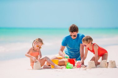 Baba ve çocuklar tropik sahilde kumdan kale yapıyorlar. Aile plaj oyuncaklarıyla oynuyor.