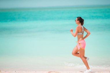 Spor kıyafetleriyle tropikal beyaz plajda egzersiz yapan zinde genç bir kadın.