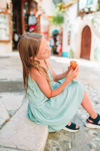 Little girl eats croissant on old streets in european village — Stock Photo, Image