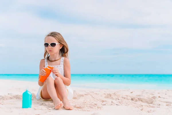 Mała dziewczynka z butelką kremu do opalania siedzi na tropikalnej plaży — Zdjęcie stockowe