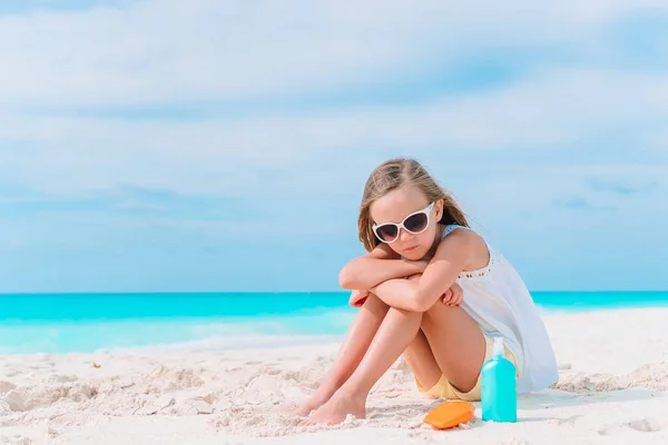 Mała dziewczynka z butelką kremu do opalania siedzi na tropikalnej plaży — Zdjęcie stockowe