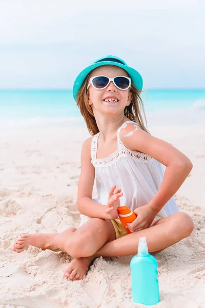 Little girl with bottle of sun cream sitting at tropical beach — Stock Photo, Image