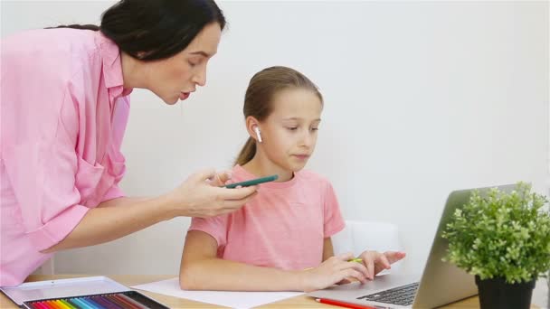 Iskoláslány ül az asztalnál a laptop és a tankönyv és a házi feladatot, míg a fiatal anya segít neki — Stock videók