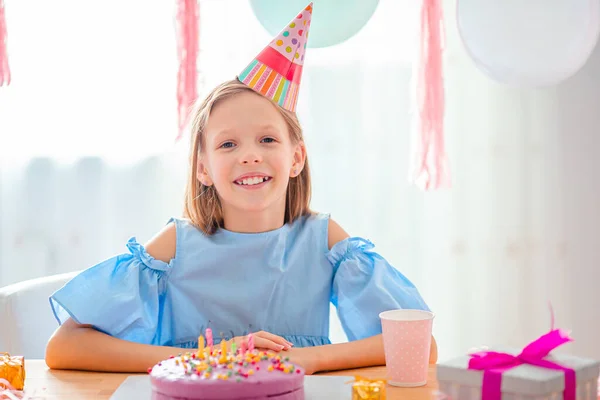 Blank meisje lacht dromerig en kijkt naar verjaardagsregenboogtaart. Feestelijke kleurrijke achtergrond met ballonnen. Verjaardagsfeest en wensenconcept. — Stockfoto