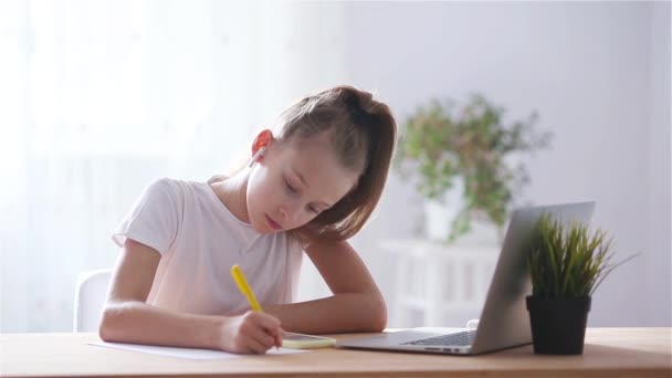 Colegiala seria sentada en la mesa con portátil y libro de texto y haciendo la tarea . — Vídeos de Stock