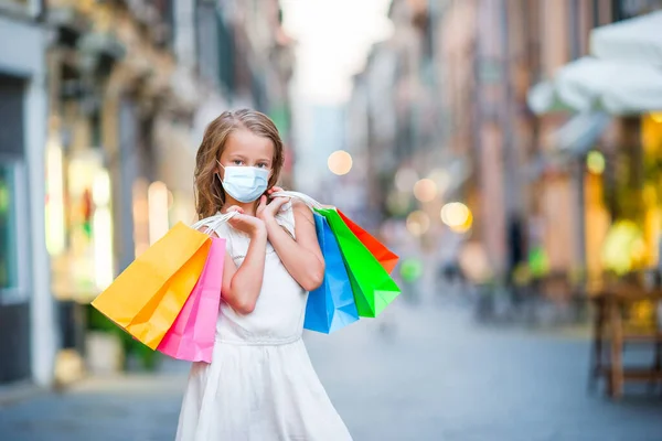 Retrato de niña adorable en máscara caminando con bolsas de compras al aire libre en la ciudad europea . — Foto de Stock