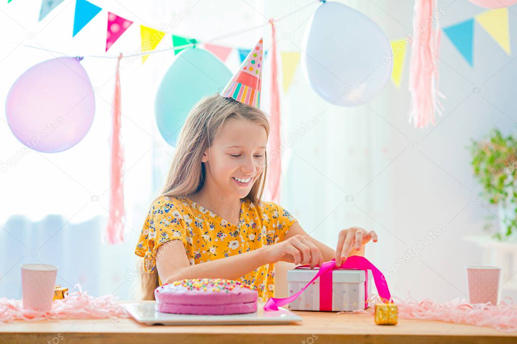 Caucasian girl is dreamily smiling and looking at birthday rainbow cake. Festive colorful background with balloons. Birthday party and wishes concept.