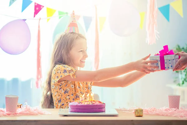 Běloška se zasněně usmívá a dívá se na narozeninový duhový dort. Slavnostní barevné pozadí s balónky. Koncept narozeninové oslavy a přání. — Stock fotografie