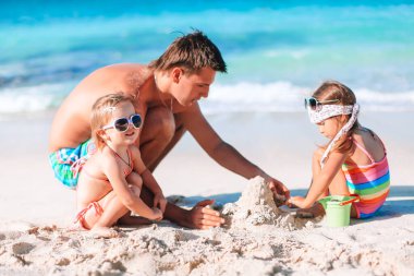 Baba ve çocuklar tropik sahilde kumdan kale yapıyorlar. Aile plaj oyuncaklarıyla oynuyor.