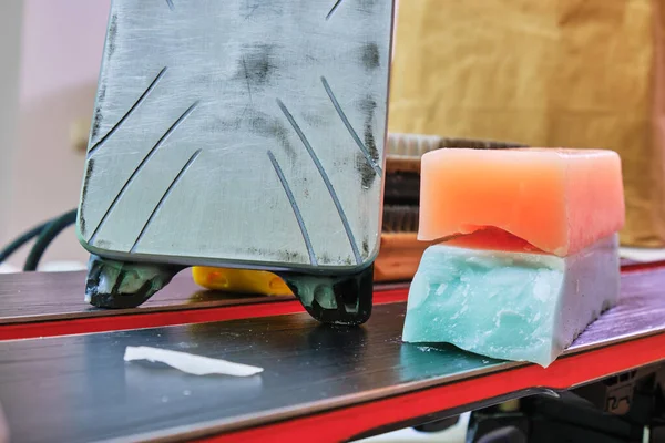 Ski wax iron and wax bars, colored orange and teal for different temperatures, laid on top of a pair of skis. Ski base tuning concept.