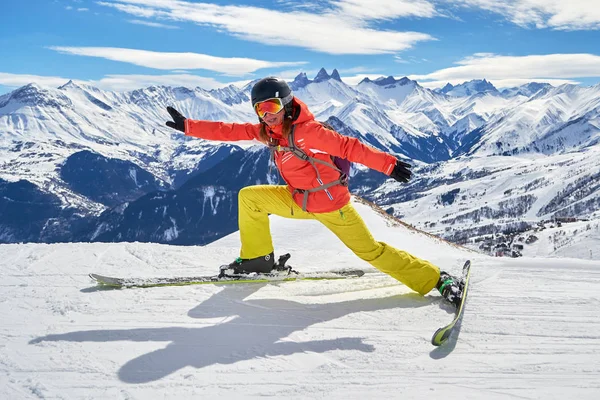 Mujer esquiadora que se extiende sobre esquís con una pose divertida, muy por encima de una pista de esquí en el dominio de esquí de Les Sybelles, Francia, en un soleado día de invierno, con los picos de Les Aiguilles d 'Arves en el fondo . — Foto de Stock
