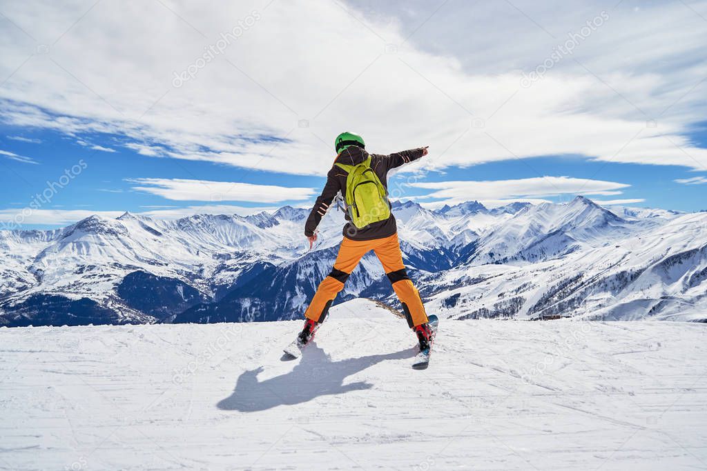 Enthusiastic skier wearing colorful clothes and a green backpack, posing on a ski slope in Les Sybelles ski resort, with French Alps peaks in the background, on a day with perfect skiing weather.