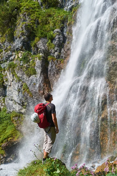 Άνδρας με λευκό κράνος στο κόκκινο σακίδιο του κοιτάζοντας το Dalfazer καταρράκτη (Dalfazer Wasserfall) πάνω από Achensee, Αυστρία, σε μια φωτεινή ηλιόλουστη μέρα. — Φωτογραφία Αρχείου