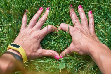 Modern teknoloji bilekliği takan bir kadın mükemmel yeşil çimlerde elleriyle kalp şeklinde bir işaret veriyor. Doğa kavramına geri bağlanılıyor.