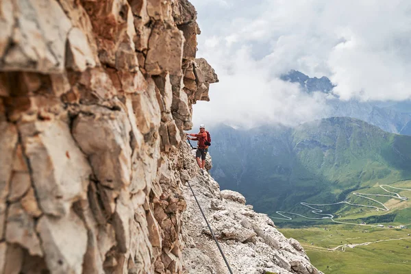 Man on via ferrata Cesare Piazzetta, Dolomites βουνά, Ιταλία, κοντά στο βράχο τοίχο, με ένα ελικοειδές δρόμο από κάτω, σε μια καλοκαιρινή μέρα με σύννεφα μαζεύονται γρήγορα πίσω. — Φωτογραφία Αρχείου
