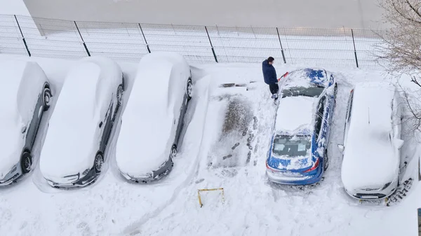 Muž čistí sníh z auta štětcem, v řadě s zaparkovaným autem pokrytým čerstvou vrstvou sněhu. Zobrazení vysokého úhlu. — Stock fotografie