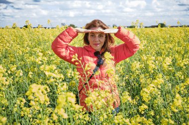Kolza tohumu tarlasında güzel bir kadın parlak güneşten gözlerini gölgeliyor. Güneş ışığını engellemek için iki elini de kullanıyor. Kameraya doğru bakıyor..