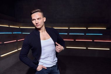 Adam beyaz tişörtlü, arka plandaki neon ışıklara yaslanmış sıradan poz veriyor.