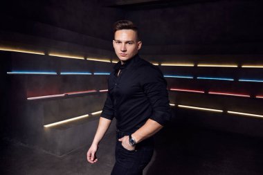 Siyah tişörtlü çekici bir adam arka planda neon ışıkları altında saat poz veriyor.