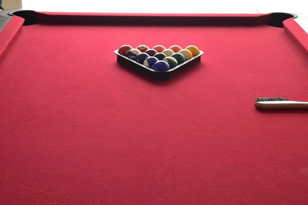 Colored billiard balls in a black ball rack with a tan wooden cleaning brush on a red felt pool table.