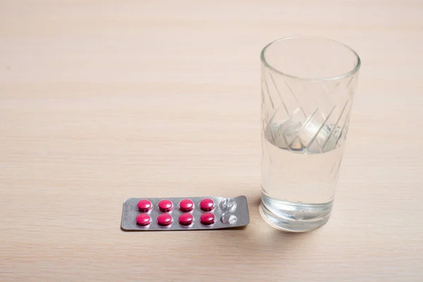 A blister with pink tablets and a glass of water stand on a light table — ストック写真