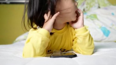 Küçük Asyalı kız yataklarında akıllı telefon izliyor. Cep telefonu bağımlısı bir çocuk.