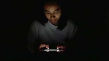 Tablet kullanan Asyalı kadın ve ekrandan gelen ışık gece yatak odasında yüzünde parlıyor..