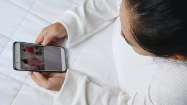 Asyalı kadın evde yatakta yatarken akıllı telefonu alışveriş için kullanıyor. Teknoloji ve rahatlama kavramı.