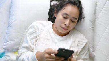 Geceleri yatakta akıllı telefon kullanan Asyalı bir kadın uykulu bir şekilde gözünü ovalıyor. Teknoloji ve Sağlık Hizmetleri kavramı.