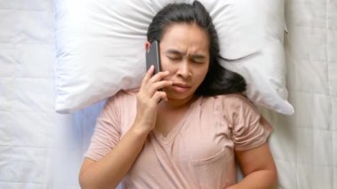 Yatakta uyurken telefona cevap vermek için uyanmak zorunda kalan kızgın Asyalı genç kadının en iyi görüntüsü. Sağlık hizmeti kavramı.