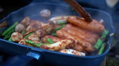 Tavada domuz eti, sosis, mantar ve baharatlı kuşkonmaz ızgara (Sichuan Pepper) ile anne eli. Asyalı aile çam ormanlarında kamp ateşinde yemek pişiriyor..