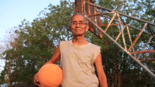 今年夏天 身穿运动服的亚洲老年人手握篮球 在室外篮球场上看着摄像机 健康生活方式和保健概念 — 图库视频影像