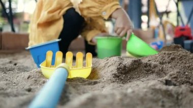 Mutlu küçük bir kız çocuğu güneşli bir yaz gününde kum havuzunda plastik oyuncaklarla oynarken eğleniyor..