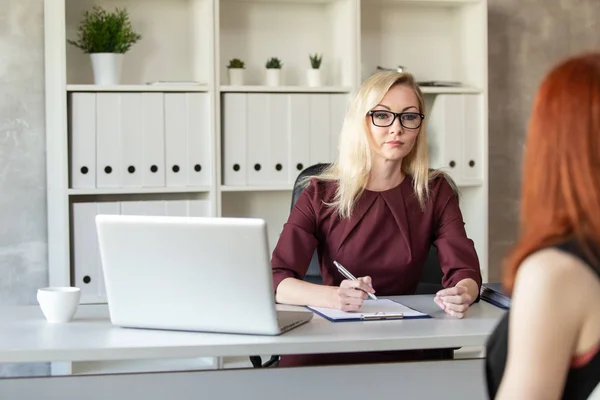 Mujer rubia en gafas se sienta detrás de la mesa de la oficina y escucha a otra mujer. Enfoque selectivo — Foto de Stock
