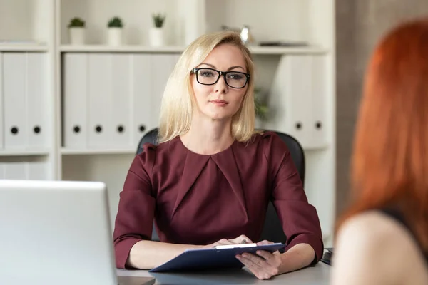 Mujer rubia en gafas se sienta detrás de la mesa de la oficina y escucha a otra mujer. Enfoque selectivo — Foto de Stock