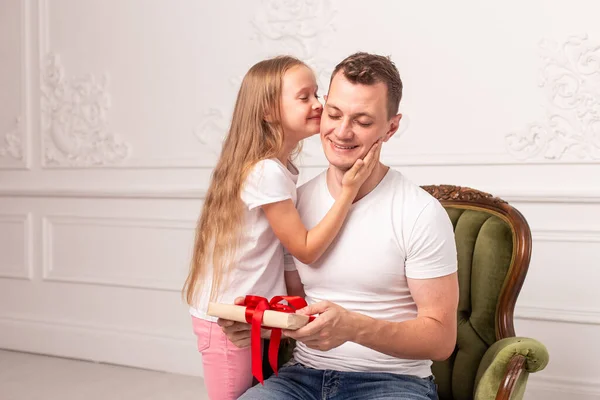 Маленькая девочка целует своего отца, который держит подарочную коробку в руке, в щеке. Селективный фокус — стоковое фото