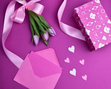 Mor arka planda pembe kurdeleli bir buket menekşe lale, pembe zarf, kalpler ve hediye kutusu, Top View